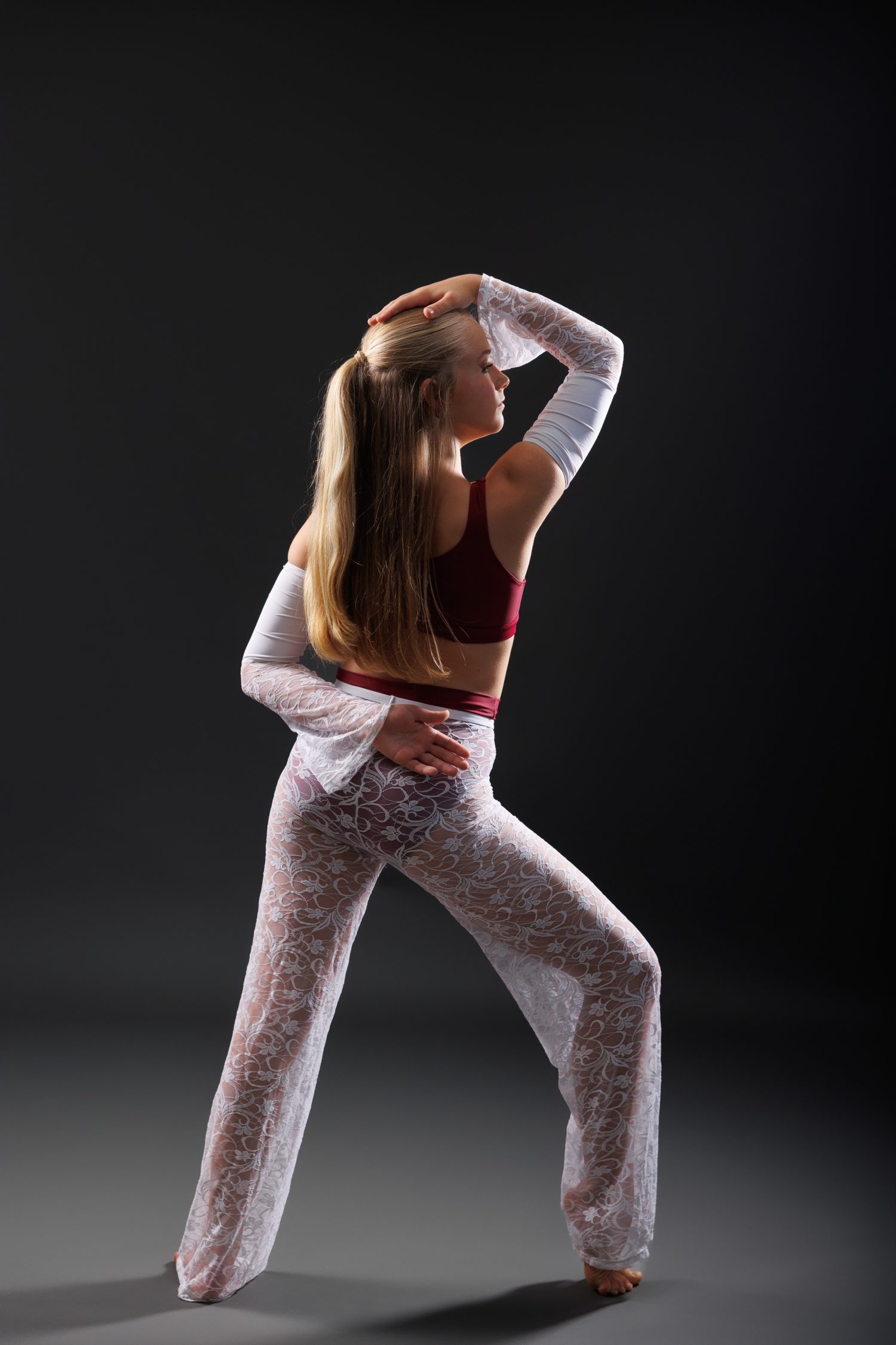 A dancer in a sheer lace costume standing barefoot with dramatic side lighting casting shadows across a grey studio floor, from a Delight Images dance photography session