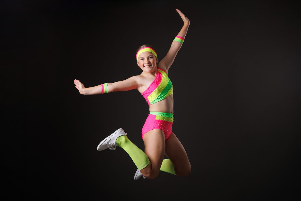 A young dancer mid-leap in a pink and green competition costume against a black studio backdrop, from a Delight Images dance portrait sessionSydney dance photographer